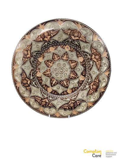Front of the round copper wall plate, showing a large mandala pattern of etched silver and copper motifs on a dark brown ground, with a small white stand behind it.